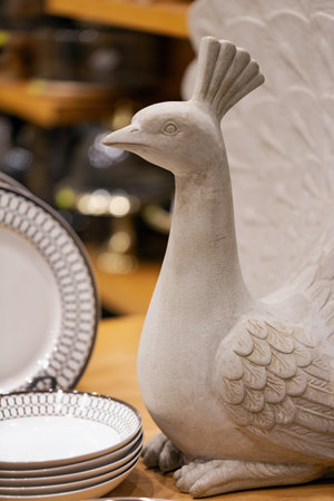 Ceramic peacock figurine in white, displayed prominently among plates. The peacock, with detailed feathers and an elegant posture, stands out. The background reveals shelves with various kitchen itemsの写真素材