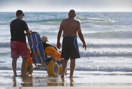 Two men are assisting an elderly man in a specialized beach wheelchair across the sandy shores. Sons taking care of their elderly parents with disabilities, spending time with them by the sea. Careの写真素材
