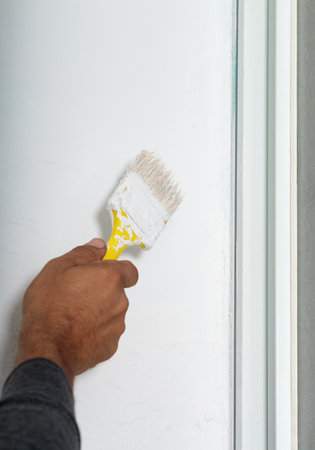 A man painting a wall white and using a paintbrush with a yellow handle. The bristles of the brush are coated in white paint. The wall appears to be near a window as evidenced by the visible frameの写真素材