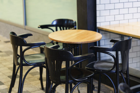 Four black chairs around a round wooden table. The chairs have a modern design with open backs. White tiles cover the wall in the background. The floor is made of square tiles. Terrace outdoorsの写真素材