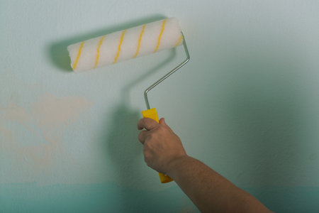 woman hand holding a new paint roller on green wall. A hand is seen applying a fresh coat of light green paint onto a wall using a yellow and white paint roller. The motion of the stroke is visibleの写真素材