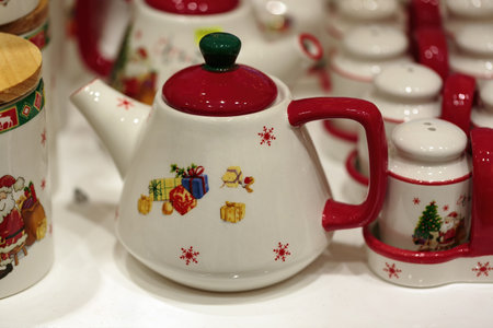 Variety of ceramic teapots and containers, all adorned with festive designs. A teapot with a white base and colorful Christmas motifs is in the foreground, include Santa, gifts, bells, and snowflakesの写真素材