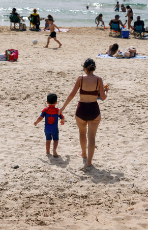 A mother and her young son are walking hand-in-hand along a sandy beach. The boy is wearing a colorful swimsuit, while the mother is dressed in a dark swimsuit. Other beachgoers are relaxing nearbyの写真素材