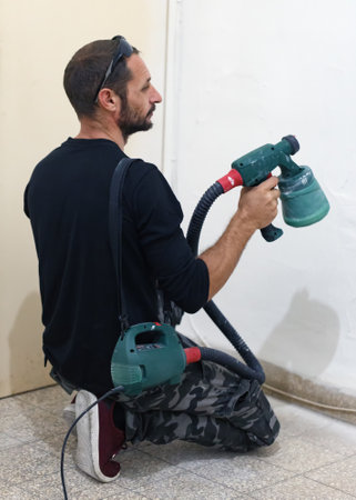 Man painting interior wall with sprayer in home during daytime. Male is kneeling and using a spray paint machine to apply paint to an interior wall. He appears focused on evenly coating the surface.の写真素材
