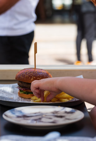Young child enjoying a burger and fries at an outdoor cafe on a sunny day. A person reaches for fries next to a delicious burger at an outdoor cafe. The background features passersby and sunny weatherの写真素材