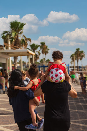 Family strolls along a picturesque beach boardwalk under blue sky with clouds. Palm trees and beachgoers in the background as the father carries one child on his shoulders, mother holds another childの写真素材