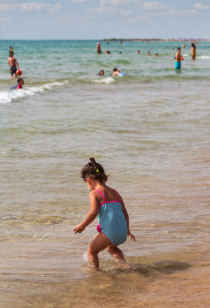 Young child wearing a blue swimsuit with pink accents is playing in the shallow waters of a sandy beach. The sunny sky and other beachgoers in the background lively and enjoyable summer day by the seaの写真素材