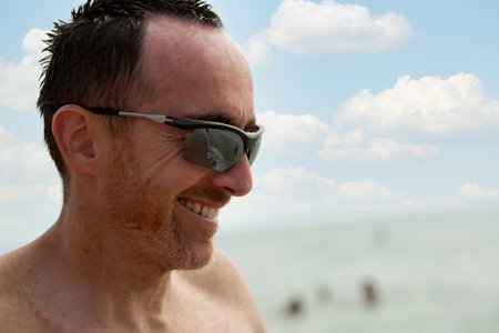A man with short dark hair and sunglasses smiles while enjoying a sunny day at the beach in the afternoon. The sky is clear with scattered clouds, and the ocean waves can be seen in the background.の写真素材