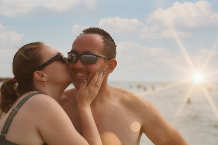 Couple enjoys a tender moment at the beach on a sunny day, with the sea waves and a bright sun in the background. They wear sunglasses and embrace with smiles, capturing affectionate summertime memoryの写真素材