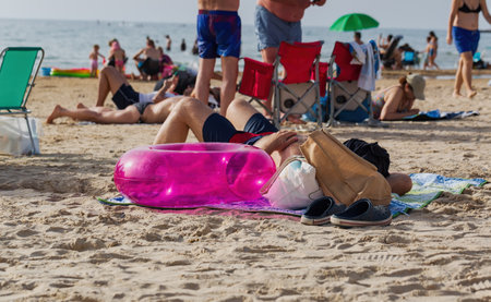 Man relaxing on beach with pink inflatable ring. A person lies on a beach towel, resting with a pink inflatable ring next to them. There are other people in the background, enjoying a day at the beachの写真素材