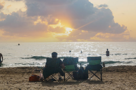 People relaxing on the beach at sun sets. Man sit in chair on a beach, watching the sunrise over the sea. Clouds adorn the sky and sun rays dance on the water surface. Beachgoers relax facing the seaの写真素材