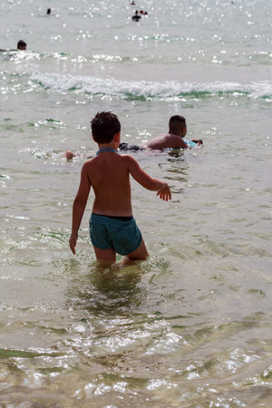 The image depicts a young child wading into the clear sea with outstretched arms. Other people swim in the distance, creating a carefree beach scene on a warm day. Summer sea and vacationers, Israelの写真素材