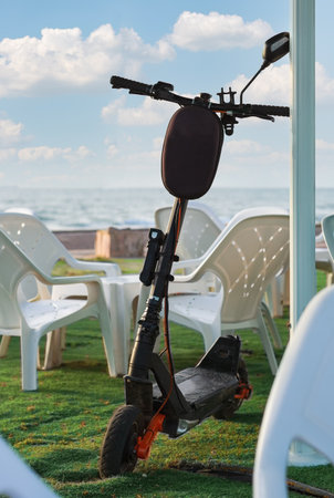 Electric scooter parked by the sea on a sunny day. Close up of black electric scooter is parked on the grass near a white table and chairs with a serene seascape and blue sky in the background.の写真素材