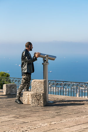 Man uses a coin operated telescope to view the Mediterranean Sea from a observation area with railing. Man is dressed in leather jacket and camouflage pants using telescope overlooking sea with boatsの写真素材