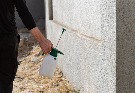 Man arm holding a white spray bottle, applying liquid to a textured wall. Spraying suggests cleaning or pest control. A person wearing a black shirt and pants is spraying wall with a spray bottle.の写真素材