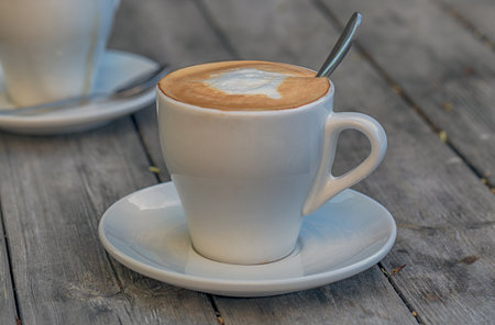 A white ceramic cup with fresh coffee and milk, a saucer, and a metal spoon on a wooden table. The coffee forms a design on top, creating an inviting scene. Cup of aroma cappuccino coffee at a cafeの写真素材