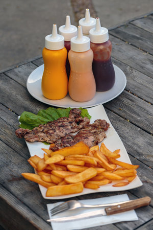 Delicious plate of grilled chicken on fresh lettuce next to golden brown French fries served with a variety of colorful sauces on a rustic wooden table. A generous serving of grilled chicken piecesの写真素材
