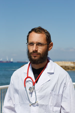 A bearded man doctor wearing glasses a white lab coat and a stethoscope with a serious look. A...
