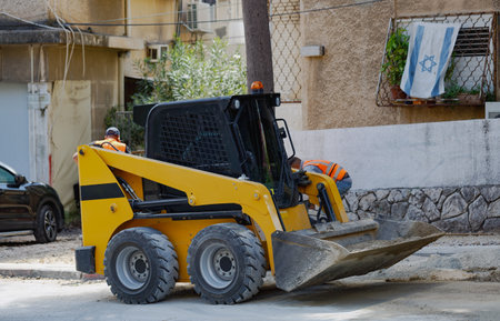 Yellow skid-steer loader with a bucket attachment in an urban setting, building a new road. Worker wearing a high-visibility vest is visible behind the small excavator at a construction site.の写真素材