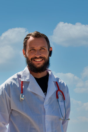 A healthcare doctor stands outdoors wearing a lab coat and a stethoscope around his neck with a happy and white smile on face. He smiles brightly against a backdrop of fluffy clouds and a blue skyの写真素材