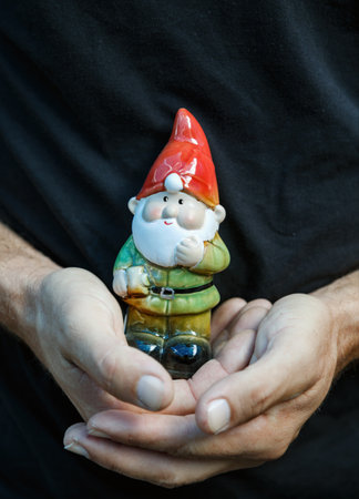 Yard gnome statue figurine in hands. A man gently holds a vibrant garden gnome, dressed in a red hat and green shirt, in front of their torso. Gnome features a thick beard and a cheerful expressionの写真素材