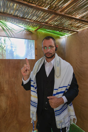 A Jewish man in a sukkah during Sukkot, wearing a black suit, shirt and a white tallit. He is raising his right index finger. The background features a wooden sukkah with a ceiling made of palm leavesの写真素材