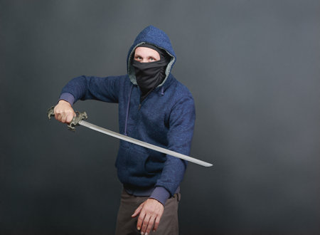 A man wearing a blue hoodie with the hood up and a black face covering, leaving only his eyes visible holds a sword, showing a dynamic pose. A person poses with a sword against a dark gray backdropの写真素材