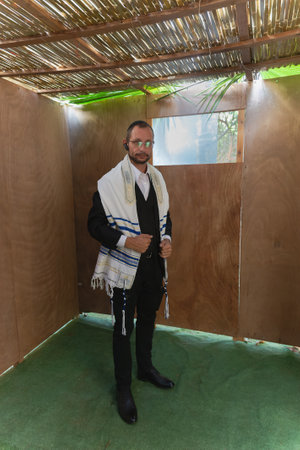 Jew man stands in a Sukkah, wearing a tallit with blue stripes and formal attire. The Sukkah has wooden walls, a palm branch roof, and grass flooring, creating a festive atmosphere. Jewish traditionsの写真素材