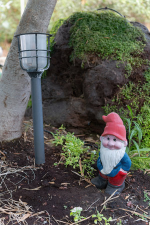 In a serene backyard, a colorful garden gnome with a red hat and blue shirt is positioned beside a modern solar lantern, moss-covered rock, while greenery surrounds the gnome. Decor features statueの写真素材