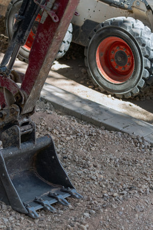 Excavator bucket and the wheels of a compact loader at a construction site with gravel and paving stones in the background, highlighting the use of industrial machinery. Construction equipment. Bucketの写真素材