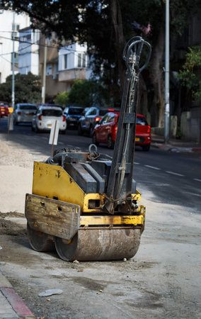 Yellow road roller parked on an urban street, used for compacting soil and asphalt. The roller shows signs of wear and is positioned near an area under roadwork with vehicles visible in the backgroundの写真素材