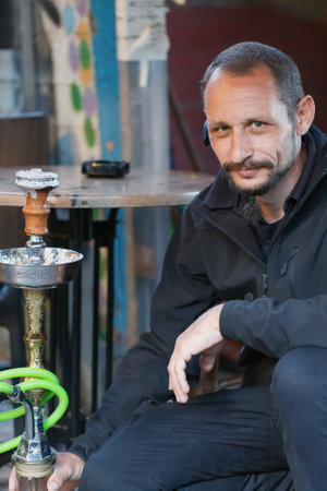A man with a beard and a slight smile is sitting indoors next to a hookah. He is wearing a dark jacket, and his posture is relaxed. The hookah and green hose, is prominently placed in the foregroundの写真素材