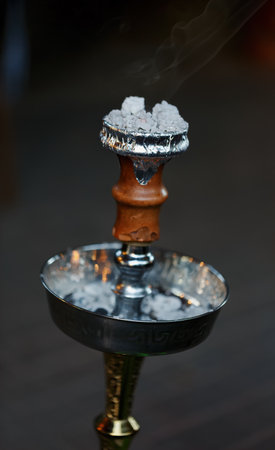 Traditional hookah bowl with burning coals on top, wrapped in foil. The bowls metallic design and wood detailing are clearly visible, and thin smoke wafts gently in the background. Hookah and shishaの写真素材