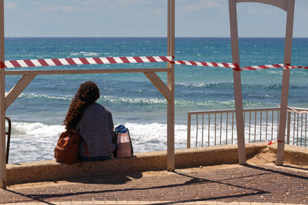 A woman with long hair sits on a stone wall by the beach, gazing at the ocean waves. The area is marked with red and white tape, indicating a restricted zone. A woman sits alone near the shore seaの写真素材