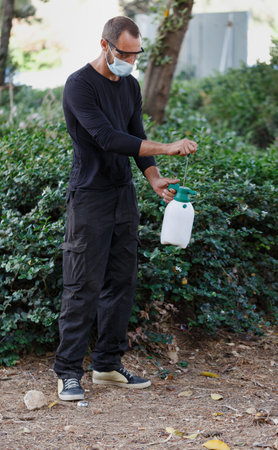 A man wearing a face mask sprays plants with a handheld garden sprayer. He is dressed casually in...