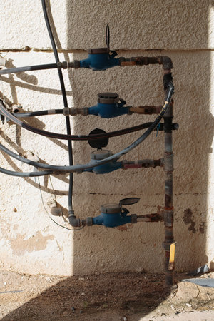 A close up shot of three blue water meters, mounted against a rough beige wall, partially in shadow. The worn pipes show signs of rust, highlighting the aging infrastructure under bright sunlightの写真素材