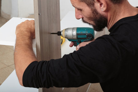 A man assembles furniture, using a cordless electric drill to secure a screw. The focus is on his careful and precise work, highlighting the DIY home improvement process. Hands on craftsmanship.の写真素材