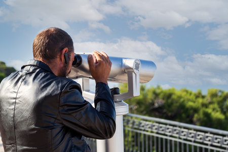 A man in a black leather jacket uses a public telescope, focused. The blurred greenery and metal...