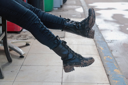 Person sitting with legs extended, wearing black leather lace-up boots covered in dried mud. The...