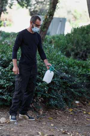 A worker in black clothing, safety goggles, and a face mask sprays plants outdoors using a handheld sprayer. His attire appears dirty, and his posture suggests focus on the task. Pesticide sprayerの写真素材