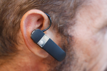 A close up of a man's ear with a black Bluetooth headset attached. Texture and facial hair are detailed, with slight redness around the ear. The focus is sharp, showing the headset's sleek design.の写真素材