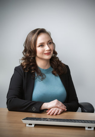 A thoughtful woman sits at a desk, wearing a teal sweater under a black blazer. Her hands are gently clasped, and a keyboard is positioned in front of her, background emphasizing professionalism.の写真素材