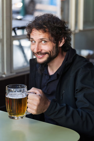 A cheerful man with a beard and curly hair smiles, sits at a table holding a beer mug. He looks directly at the camera with a slight smirk. He wears a black jacket. Happy man enjoying a cold beerの写真素材