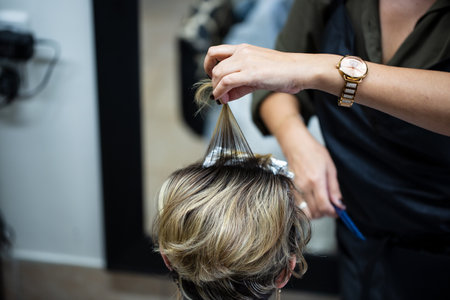 In salon, hairstylist carefully applies highlights to a section of hair, bleaching it to a blonde shade. The stylist's hands skillfully hold the hair and comb, with the backgroundの写真素材