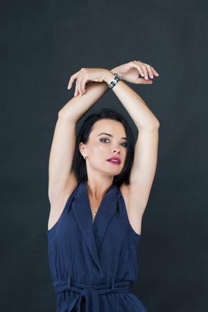 A young woman stands confidently against a dark backdrop, arms raised and crossed above her head. She wears a sleeveless dark blue dress with a belt and a wristwatch, presentingの写真素材