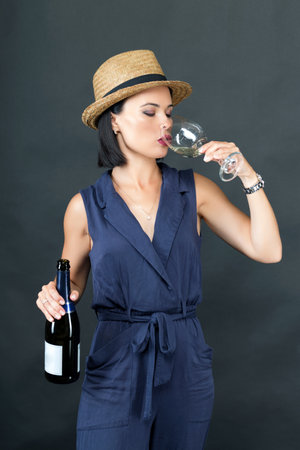 Pretty and young woman drinking wine while holding a wine bottle on a gray background. In a hand a glass with champagne. woman drinks from a glass. Portrait of youngの写真素材