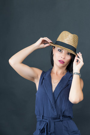 A woman in a navy blue sleeveless outfit adjusts a straw hat. The plain dark background highlights her elegant and stylish look. Portrait of a beautiful young brunette woman in ahの写真素材