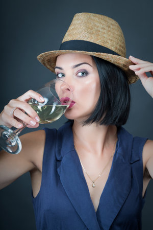Beautiful young model in a blue dress and hat drinking wine on gray background. Girl drinks sparkling wine to celebrate the new year.の写真素材