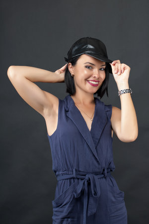 A stylish young woman with short black hair wears a navy blue sleeveless jumpsuit and a black fedora hat. She smiles confidently while playfully adjusting her hat with both handsの写真素材