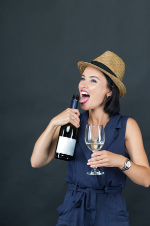 A cheerful stylish beautiful young woman enjoys champagne, wine. She playfully licks a wine bottle with an open mouth, expressing excitement.の写真素材